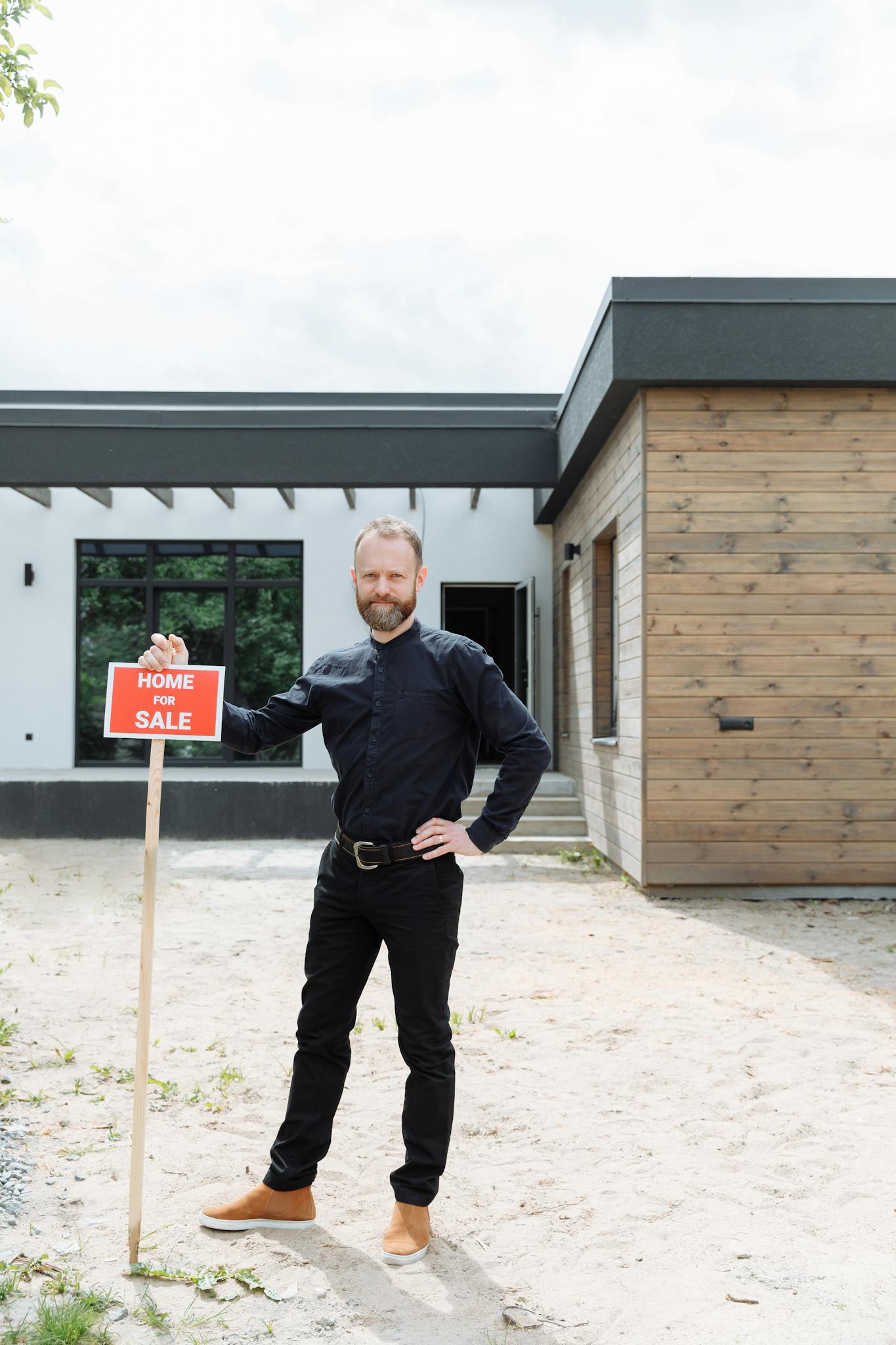 A real estate agent stands outdoors holding a 'Home for Sale' sign in front of a modern house.