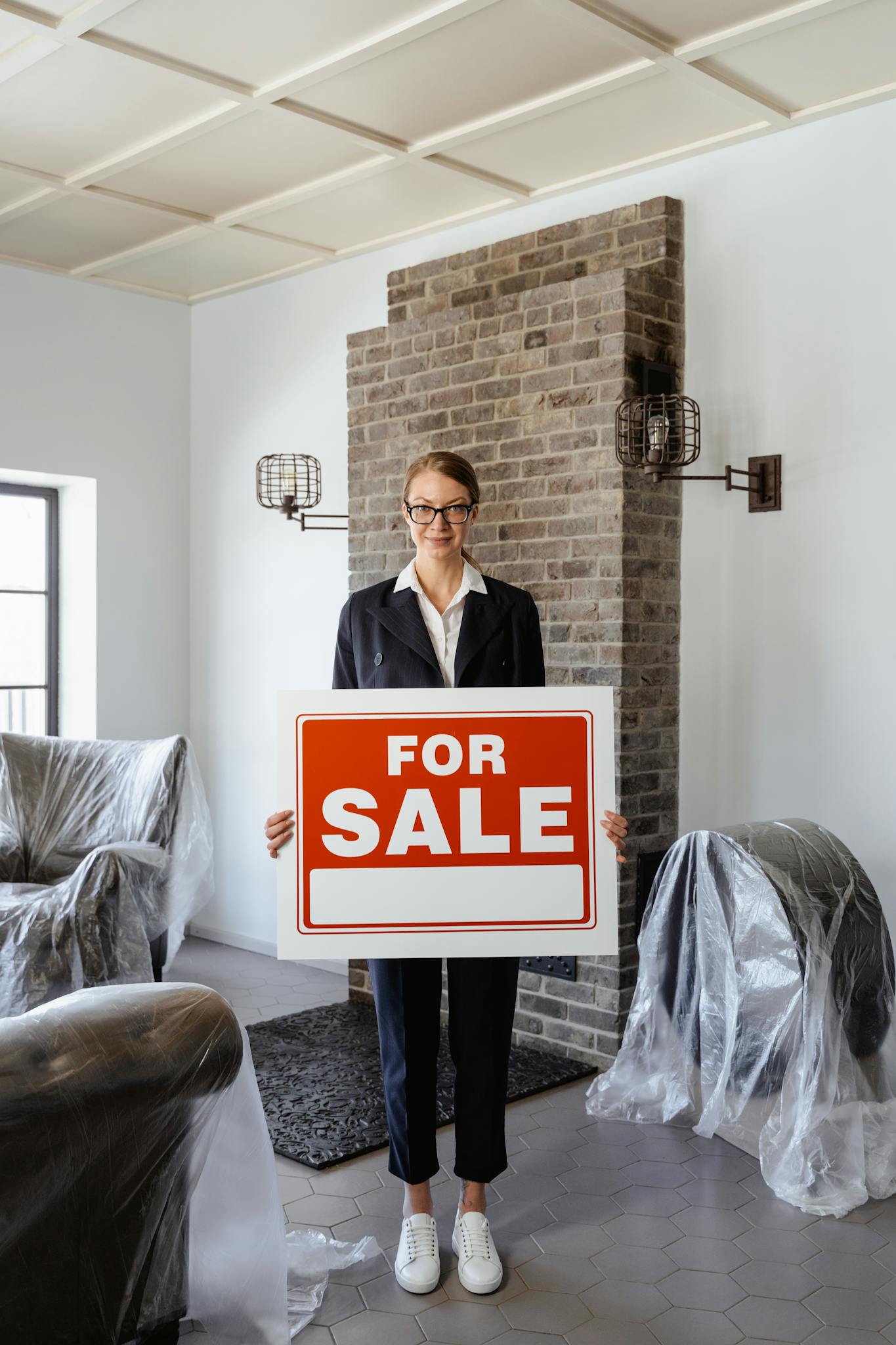 Female real estate agent indoors holding a 'For Sale' sign, showcasing property listing.